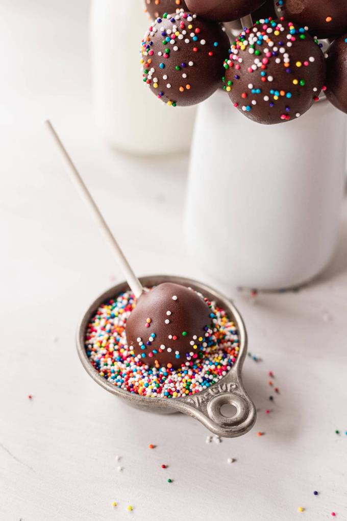 A chocolate cake pop being rolled in sprinkles. More cake pops are in a vase in the background.