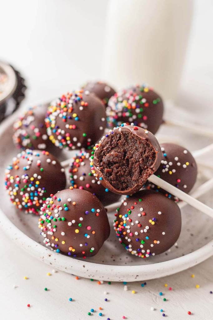 Chocolate cake pops piled on a white speckled plate. One cake pop has a bite missing. 