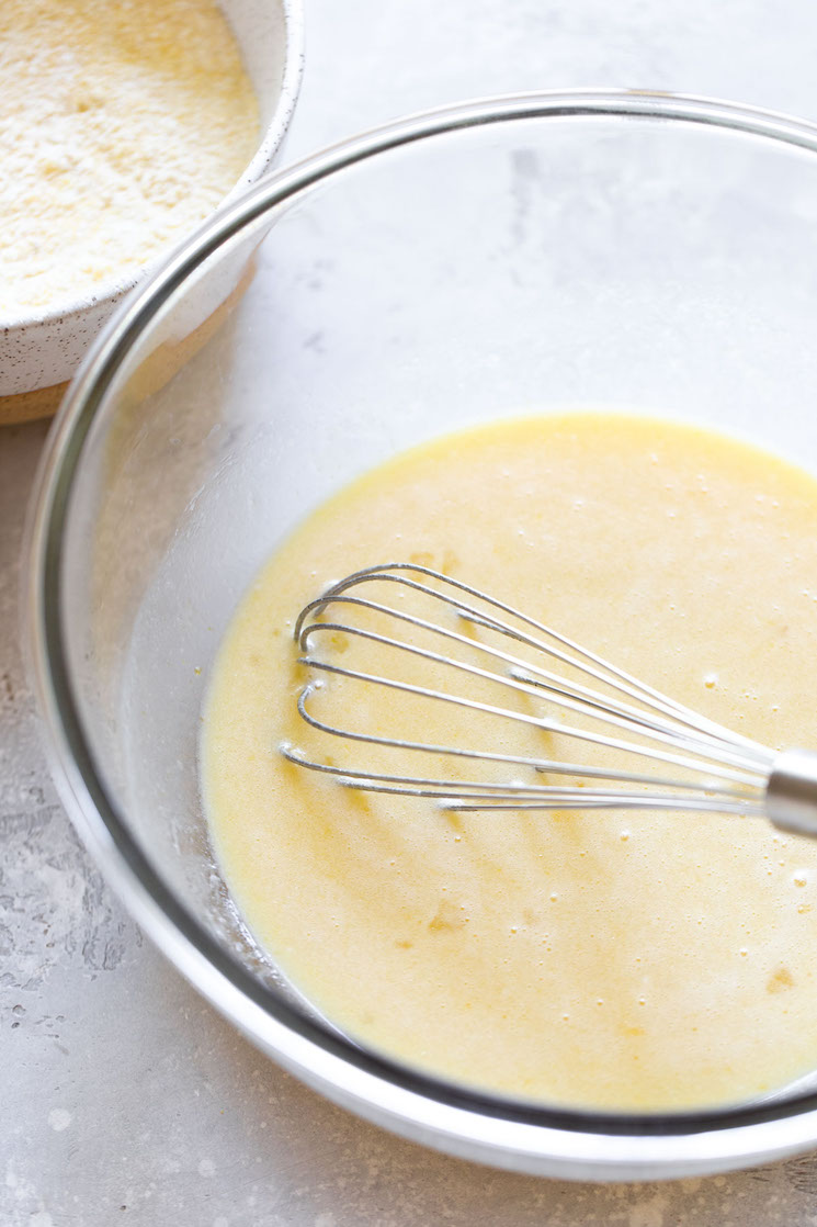 The wet ingredients needed to make cornbread being mixed in a glass bowl with the dry ingredients in the background.