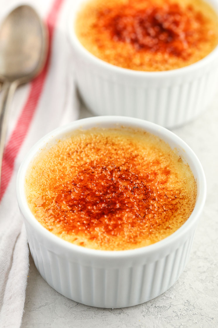 Two white ramekins filled with homemade crème brûlée and a white napkin and two spoons on the side.