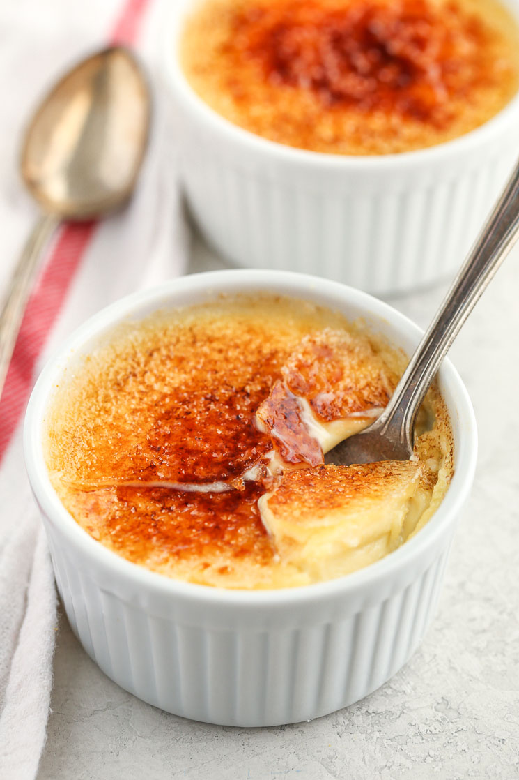 A close-up of a white ramekin filled with crème brûlée and a spoon inside of it along with another crème brûlée and white napkin.