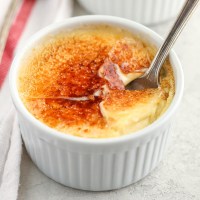 A close-up picture of a white ramekin filled with crème brûlée and a spoon inside of it.