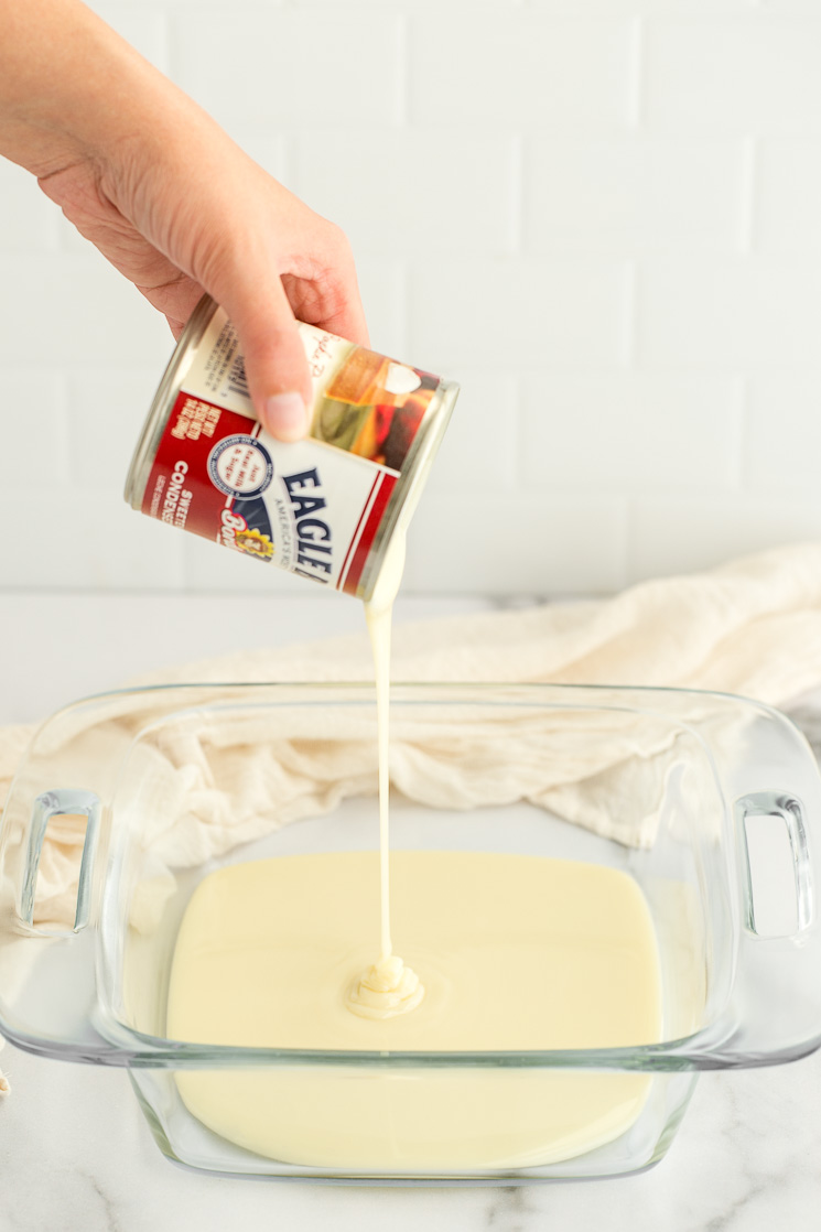 A can of sweetened condensed milk being poured into a clear baking dish.