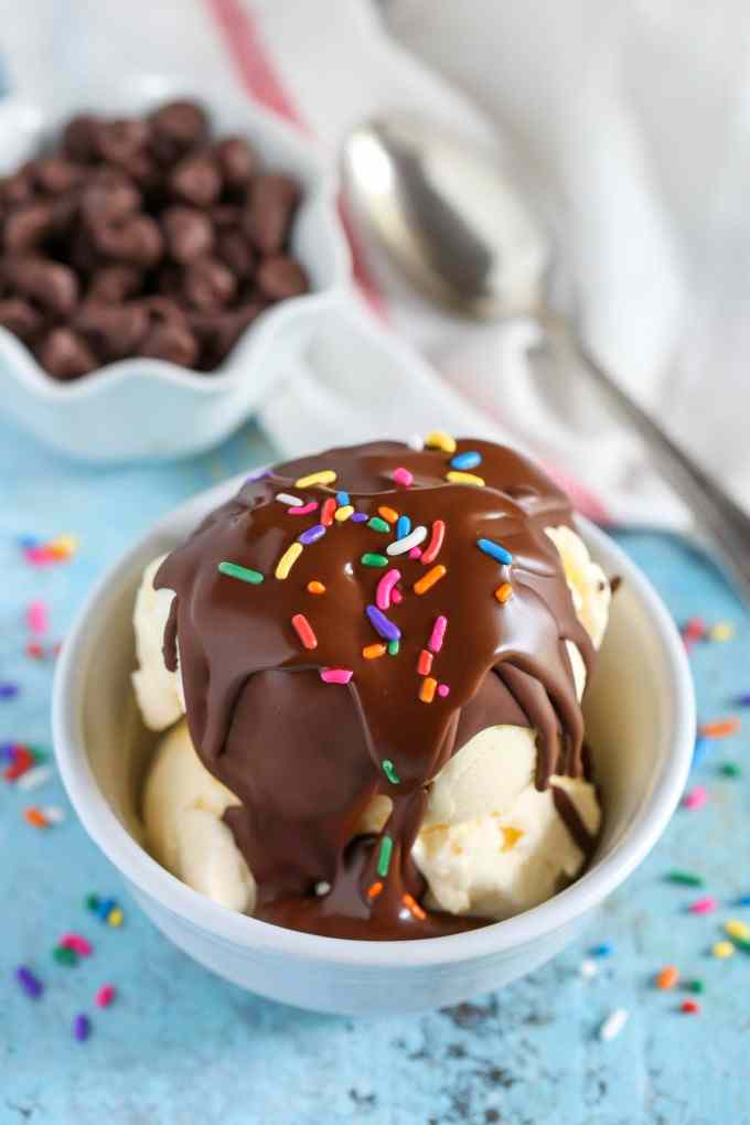 A small dish of vanilla ice cream topped with magic shell and sprinkles. A dish of chocolate chips and a spoon rest in the background. 