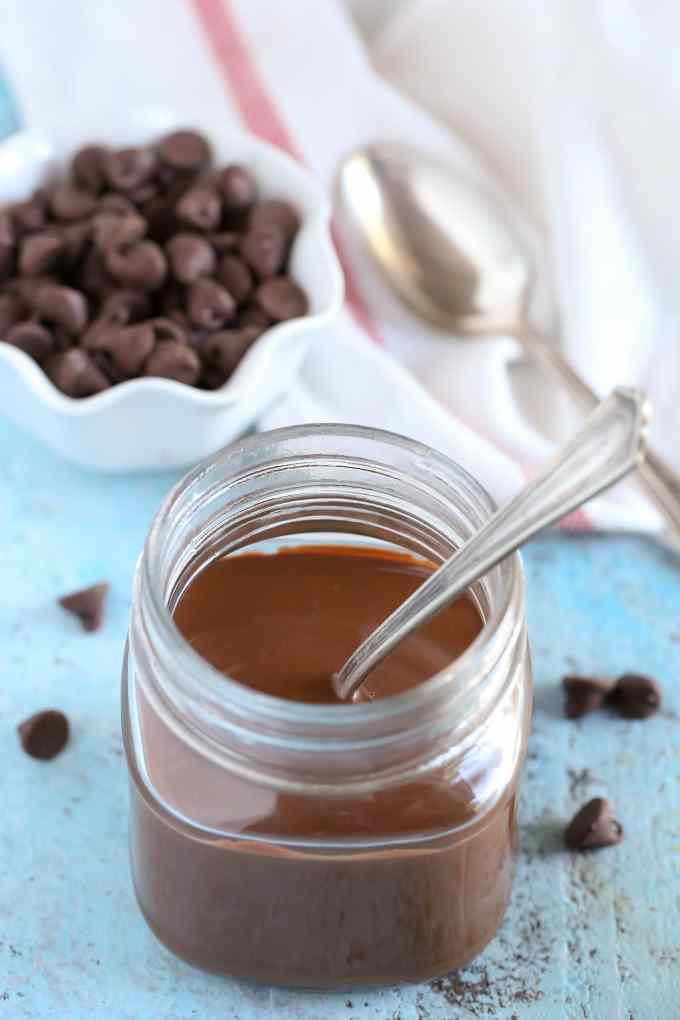 Homemade magic shell in a glass jar. A spoon is sticking out of the jar. A small dish of chocolate chips, a tea towel, and a spoon rest in the background. 