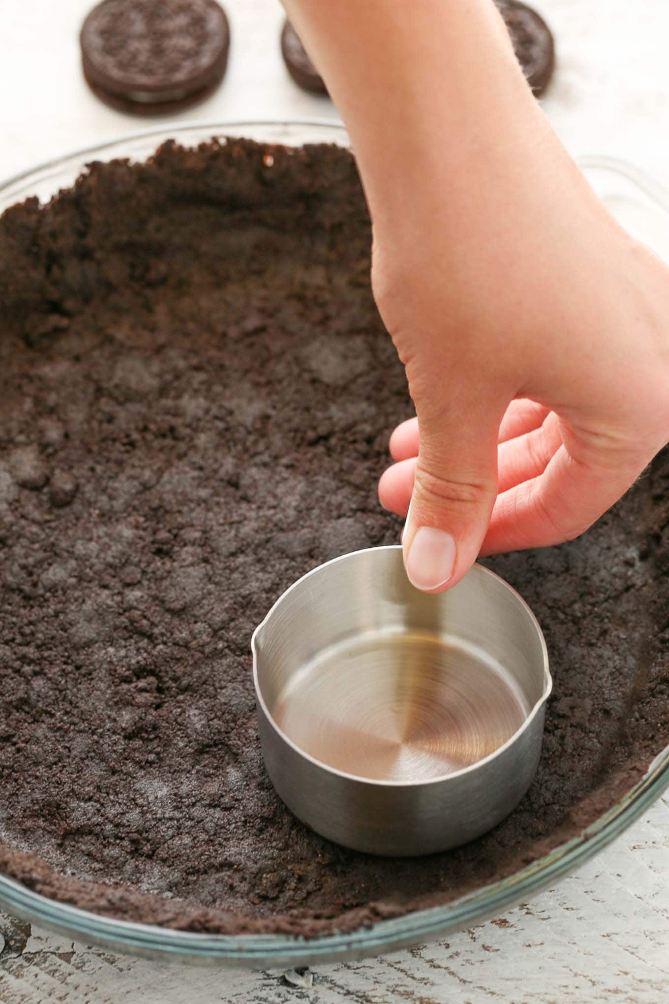 Process shot showing a measuring cup being used to pack down an Oreo cookie pie crust. 