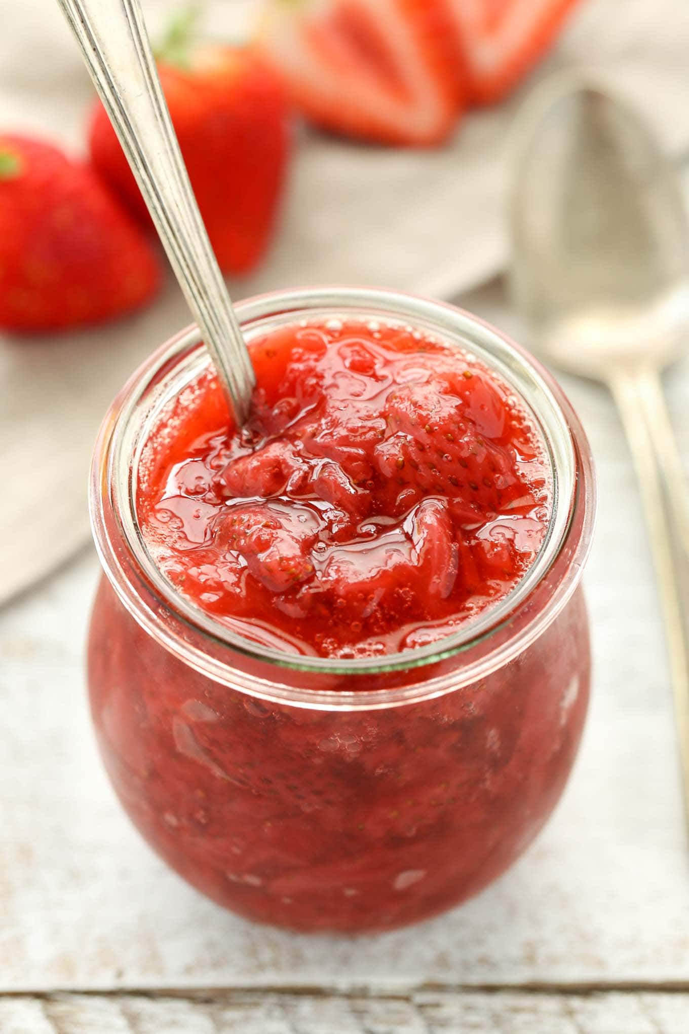 Homemade strawberry sauce in a glass jar with a spoon sticking out. Fresh strawberries rest in the background. 