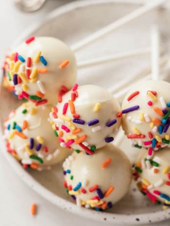 Six vanilla cake pops topped with sprinkles on a white plate.