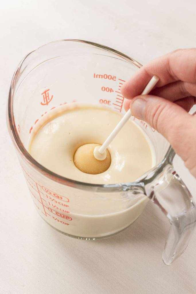 A vanilla cake pop being dunked into a glass of melted white chocolate. 