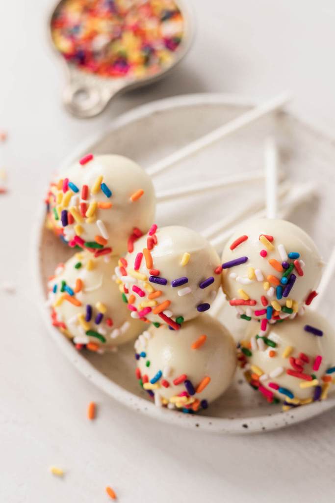 Six birthday cake pops resting on top of each other on a small white plate. Sprinkles in a measuring cup rest in the background. 