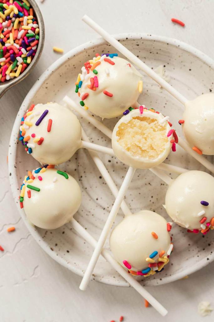 An overhead view of birthday cake pops on a small speckled plate. One cake pop has a bite missing. 