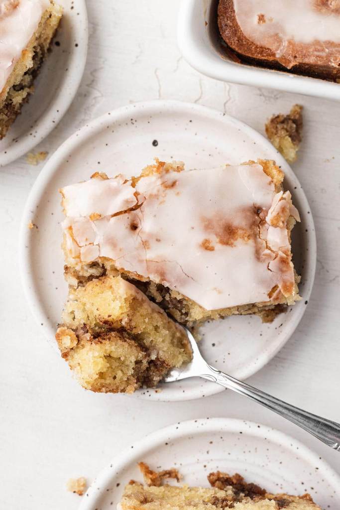 An overhead view of a slice of iced honey bun cake. A fork has speared a bite of cake and rests on its side on the plate. 