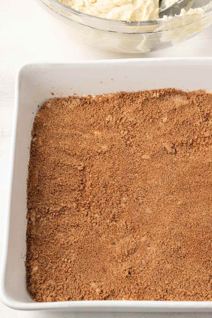 A process shot of cinnamon sugar filling in a white baking dish. A bowl of cake batter is in the background. 