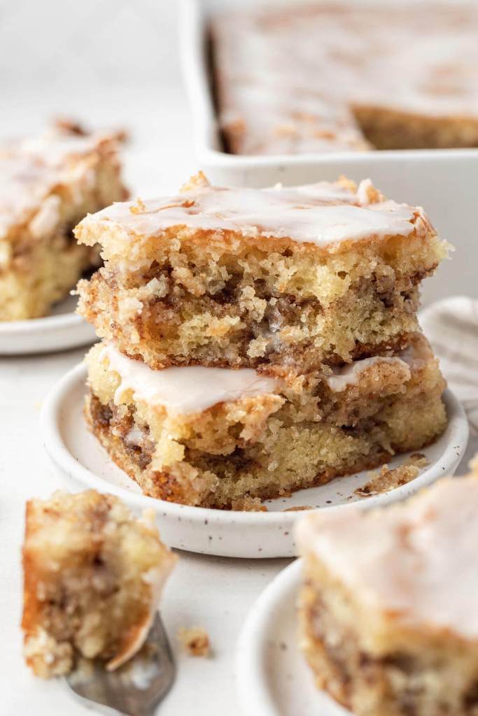 Two slices of honey bun cake, stacked on a white speckled dessert plate. Additional cake slices and a baking pan rest in the foreground and background. 