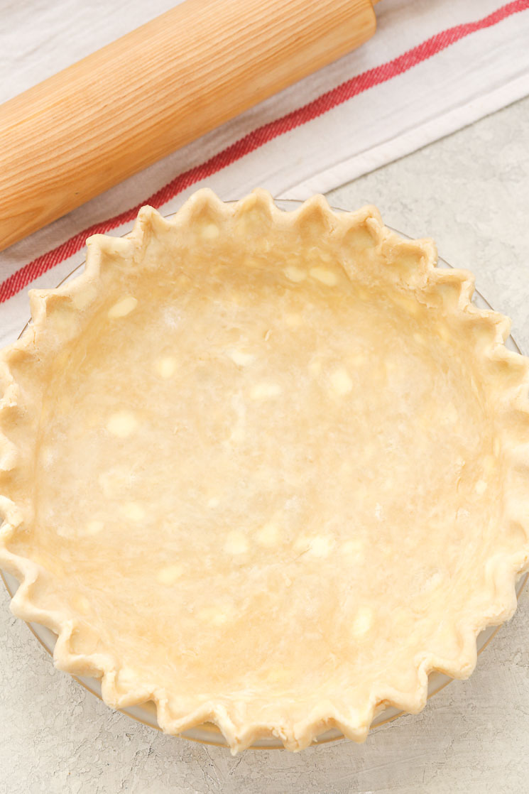 A pie crust in a glass pie dish that's ready to be blind baked. A wood rolling pin and striped towel are next to the crust. 