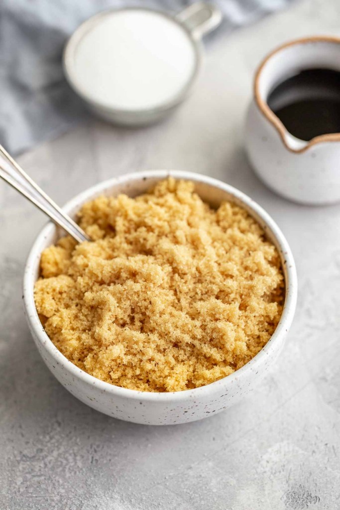 A homemade brown sugar substitute in a white bowl with a spoon sticking out. A small jug of molasses rests nearby. 