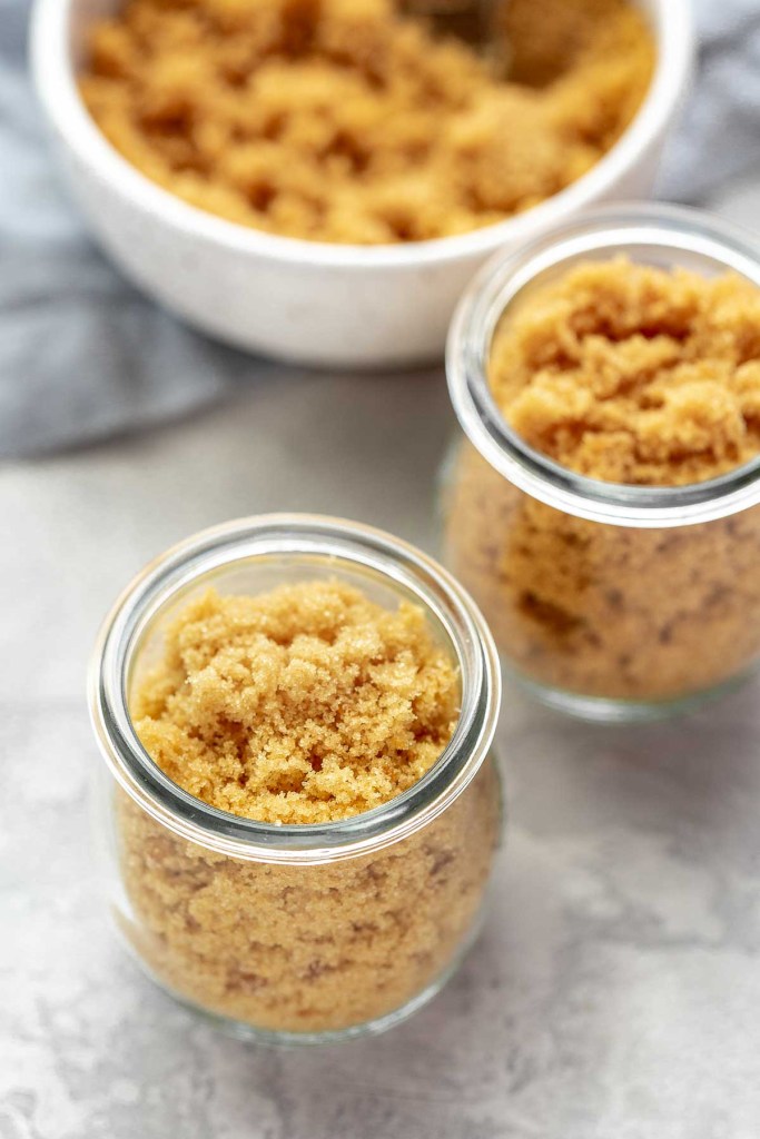 Two glass jars full of a homemade brown sugar substitute. A bowl of brown sugar rests in the background. 