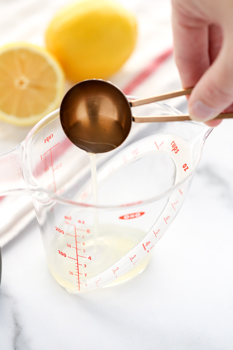 A clear measuring cup with one tablespoon fresh lemon juice. A white napkin and lemons in the background.