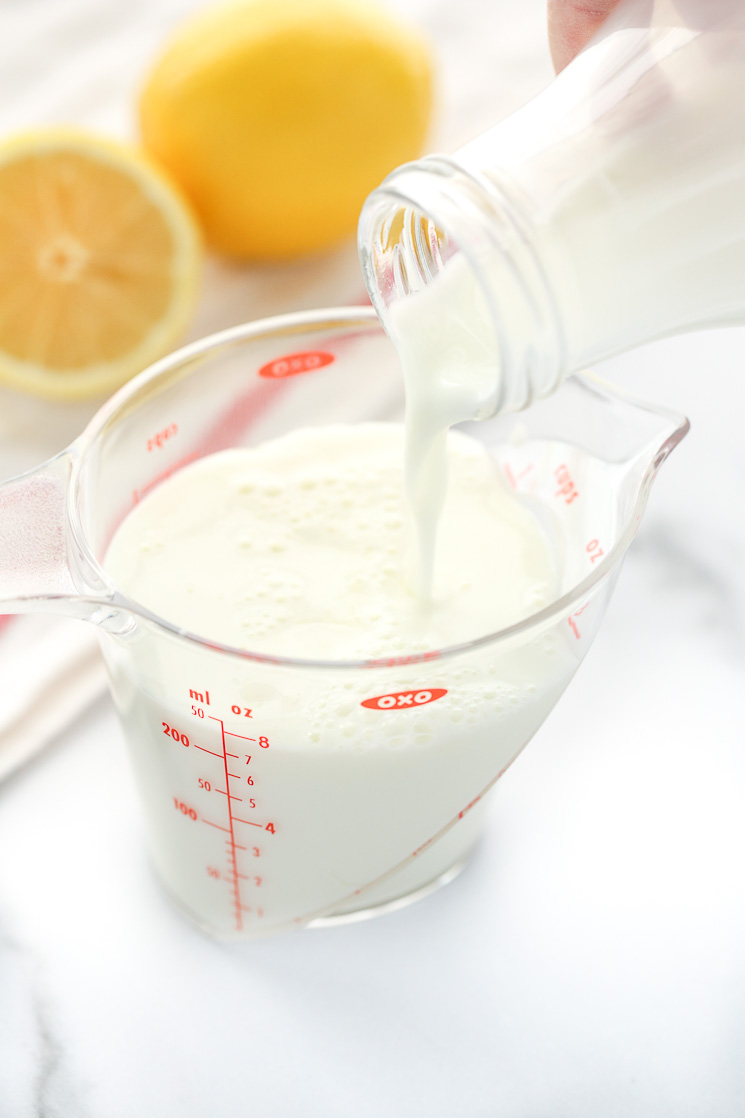 A clear measuring cup being filled with milk. A white napkin and lemons in the background.