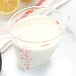 A clear measuring cup filled with homemade buttermilk along with a white napkin and lemons in the background.