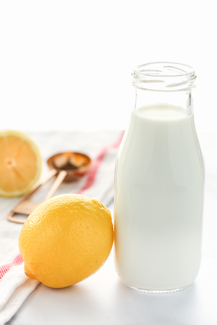 An image of a clear glass measuring cup filled with milk and a lemon beside it. A white napkin, copper measuring spoon, and sliced lemon in the background.