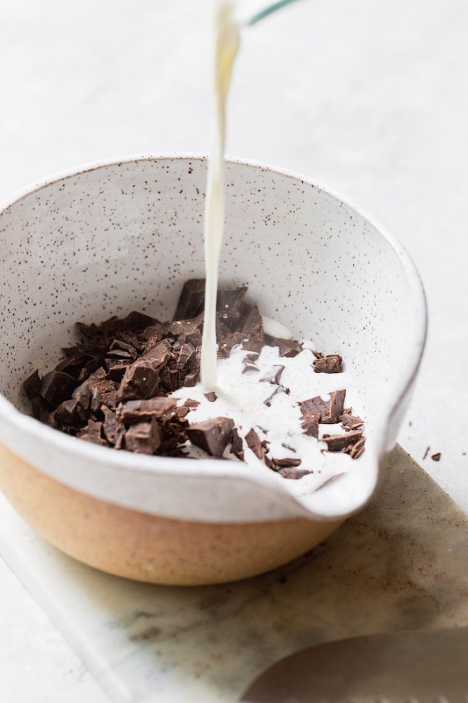 A small bowl holding chopped chocolate and warm heavy cream being poured over the top of it.