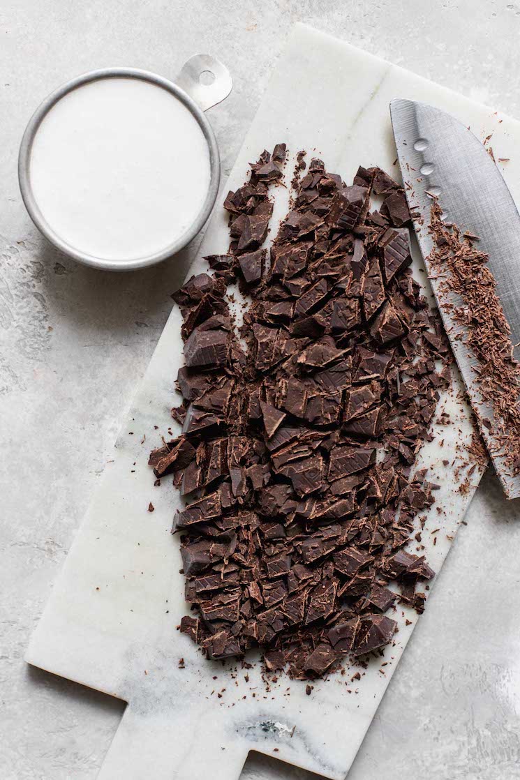 A marble cutting board covered with chopped chocolate and a small cup filled with heavy cream beside it.