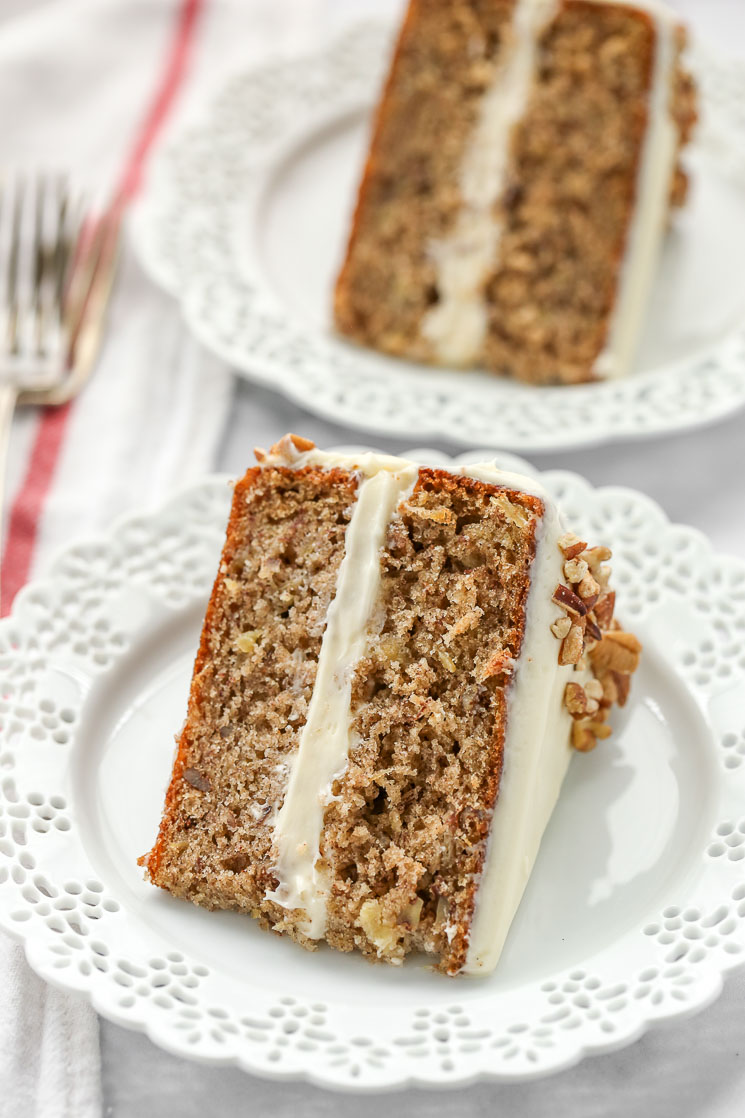 A slice of hummingbird cake on a decorative white plate with another slice and a fork in the background. 