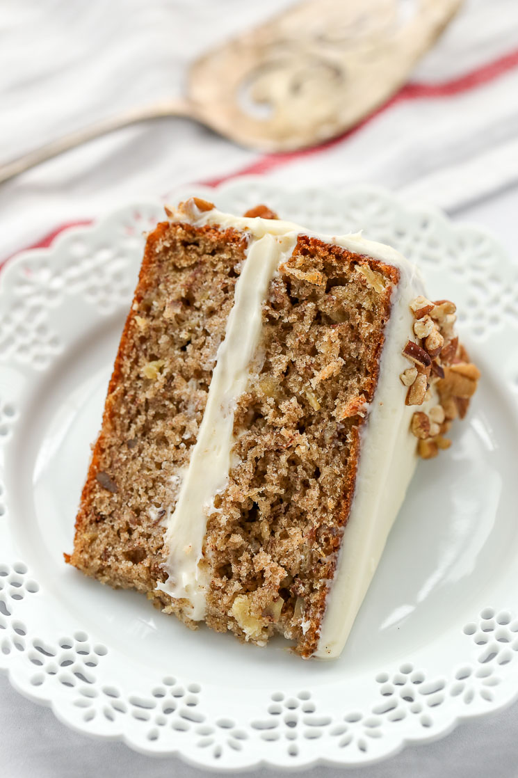 A slice of hummingbird cake on a decorative white plate with a cake server in the background.