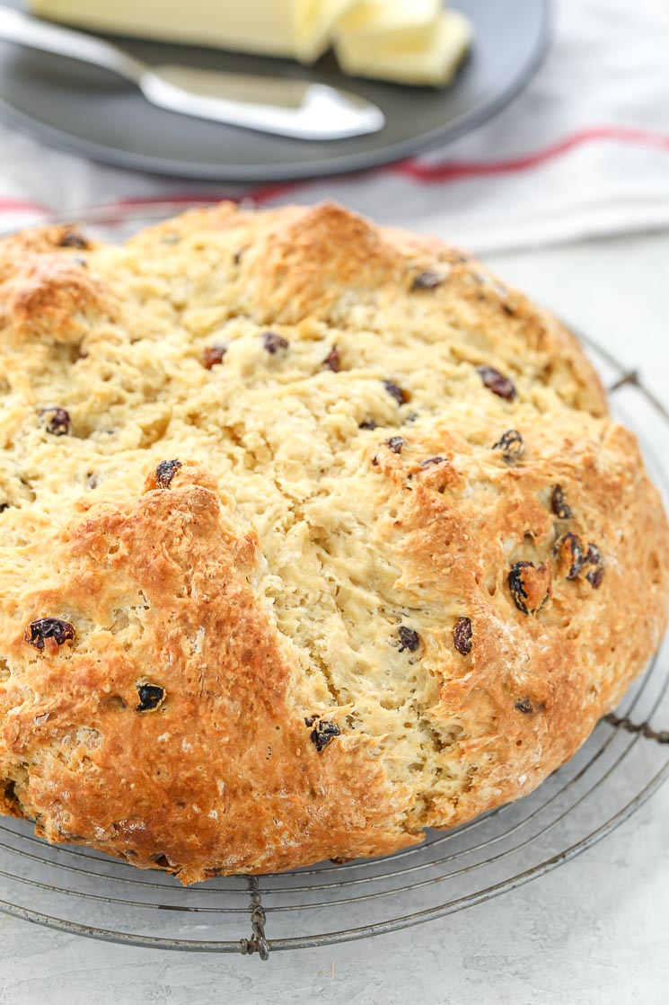 A round loaf of Irish Soda Bread with raisins on top of an antique round cooling rack. There is a black plate with sliced butter in the background. 