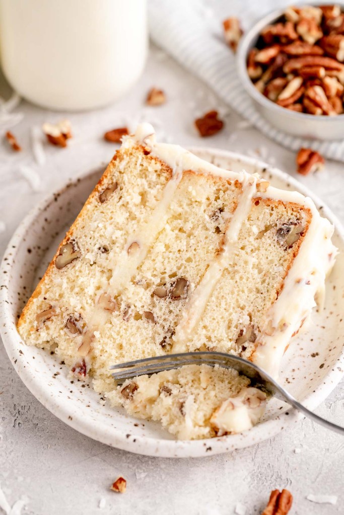 A slice of Italian cream cake on a white speckled plate. Pecans and coconut are scattered around the plate.