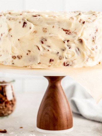 An Italian cream cake on a wooden cake stand with a marble top. A jar of chopped pecans and two milk jugs rest in the background.