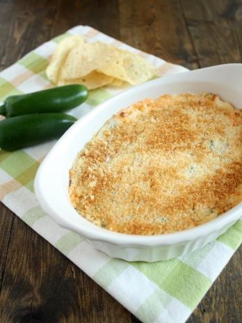 Jalapeno cream cheese dip in a small baking dish on a checkered towel. Two jalapenos and some tortilla chips rest on the towel as well.