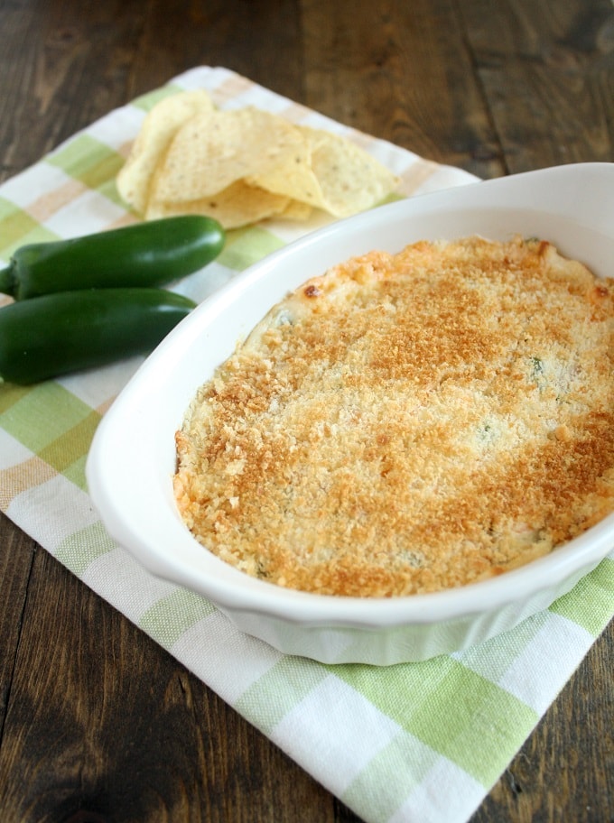 Jalapeno cream cheese dip in a small baking dish on a checkered towel. Two jalapenos and some tortilla chips rest on the towel as well. 