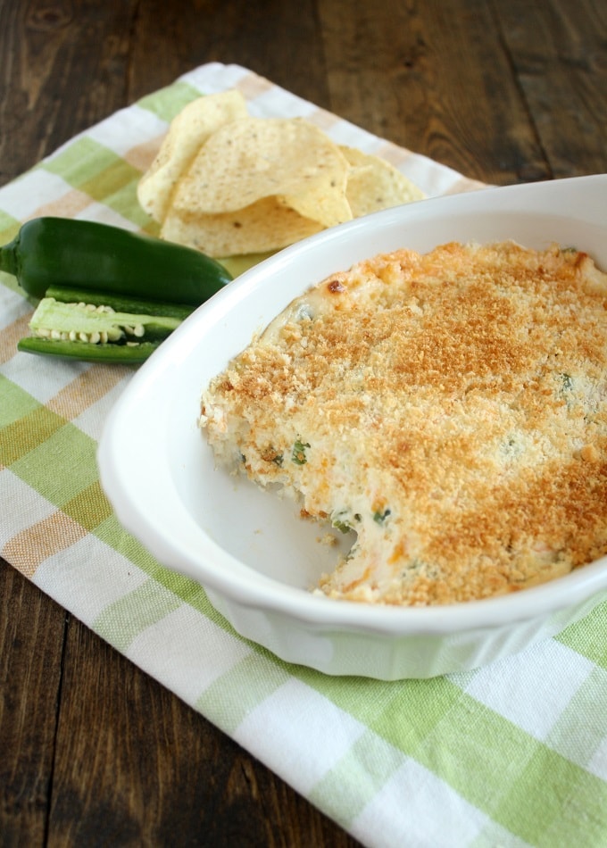 Jalapeno cream cheese dip in a small baking dish on a checkered towel. Two jalapenos and some tortilla chips rest on the towel as well. A portion has been scooped out of the dip. 