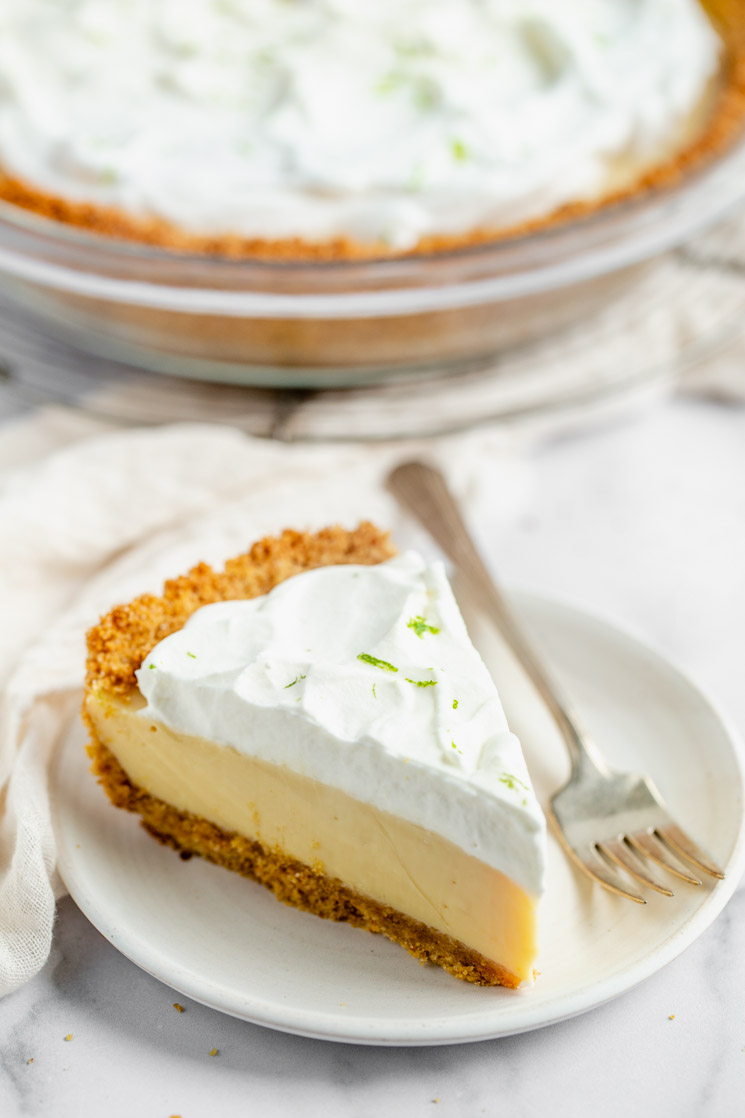 A slice of key lime pie on a white plate with the rest of the pie in the background.