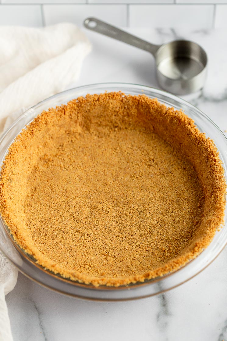 A baked graham cracker crust in a pie plate with a measuring cup behind it.