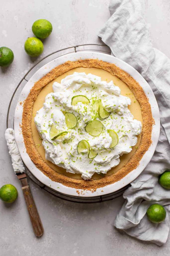 An overhead view of a traditional key lime pie, topped with whipped cream and lime slices. Key limes are scattered around the pie. 