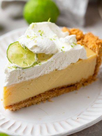 A slice of key lime pie on a white dessert plate. Key limes are scattered in the background.