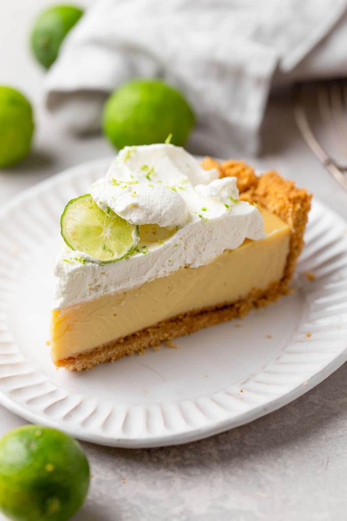 A slice of key lime pie on a white dessert plate. Key limes are scattered in the background. 
