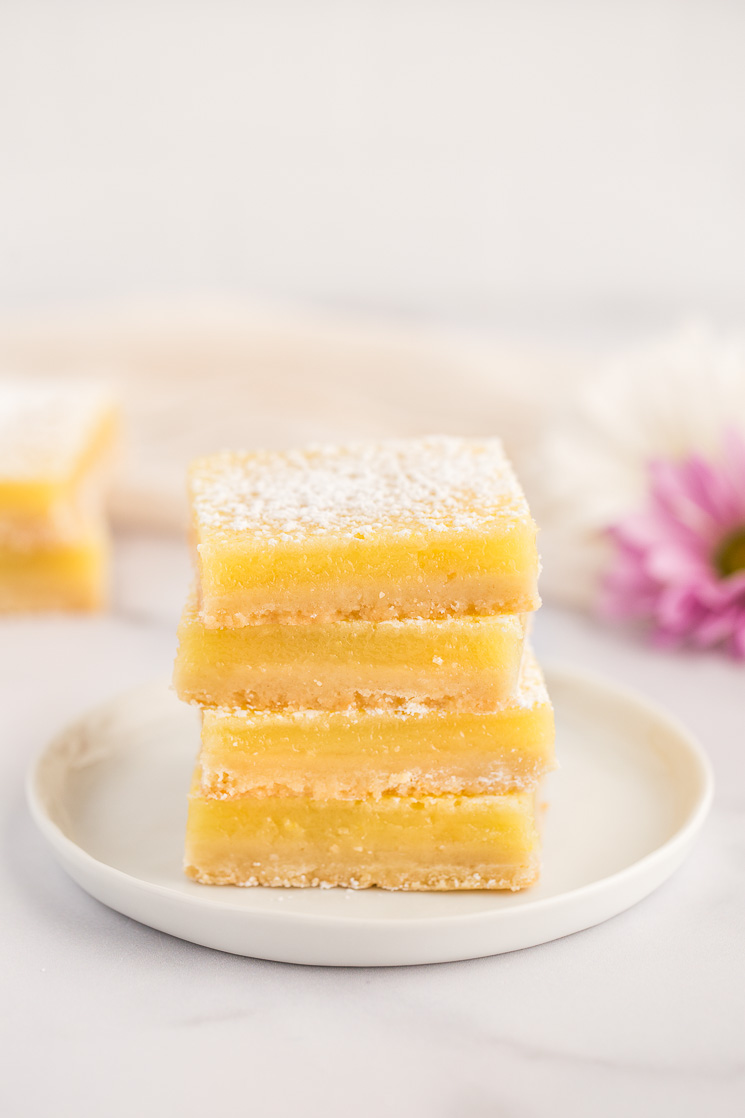 A stack of lemon bars on a white plate with more lemon bars and flowers in the background.