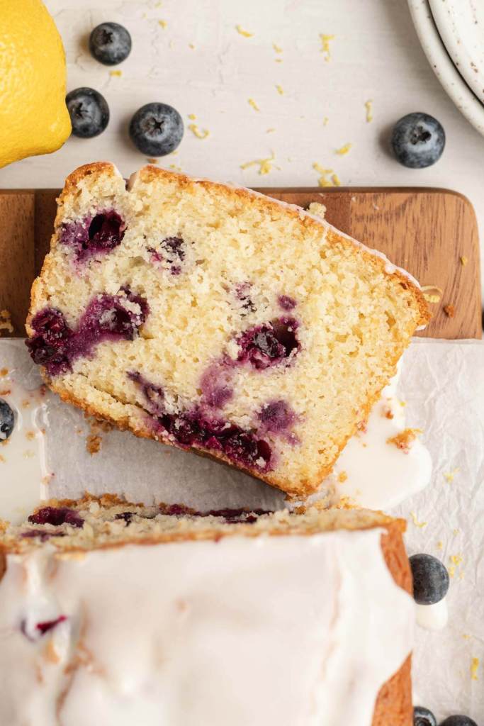 An overhead view of a lemon blueberry bread on a wood cutting board, with a slice lying on its side. 