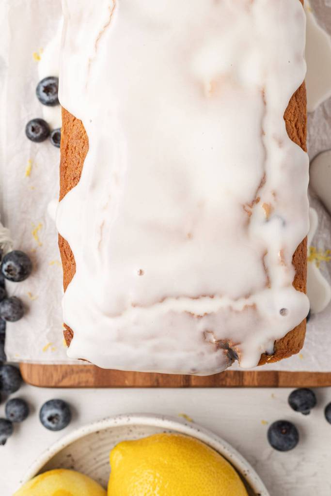 An overhead view of a loaf of lemon blueberry bread with lemon icing on a wooden board surrounded by fresh blueberries. 