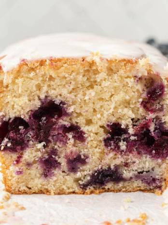 A straight on view of a loaf of lemon blueberry bread on a piece of parchment paper. The bread has been sliced to show the interior texture. A bowl of blueberries rests in the background.