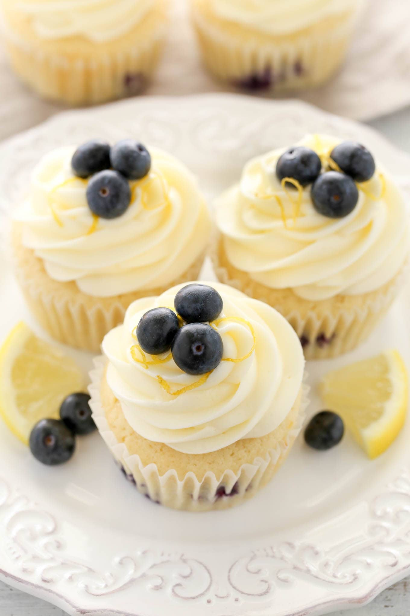 Three frosted lemon blueberry cupcakes on a white dessert plate. The plate is garnished with lemon wedges and fresh berries. 