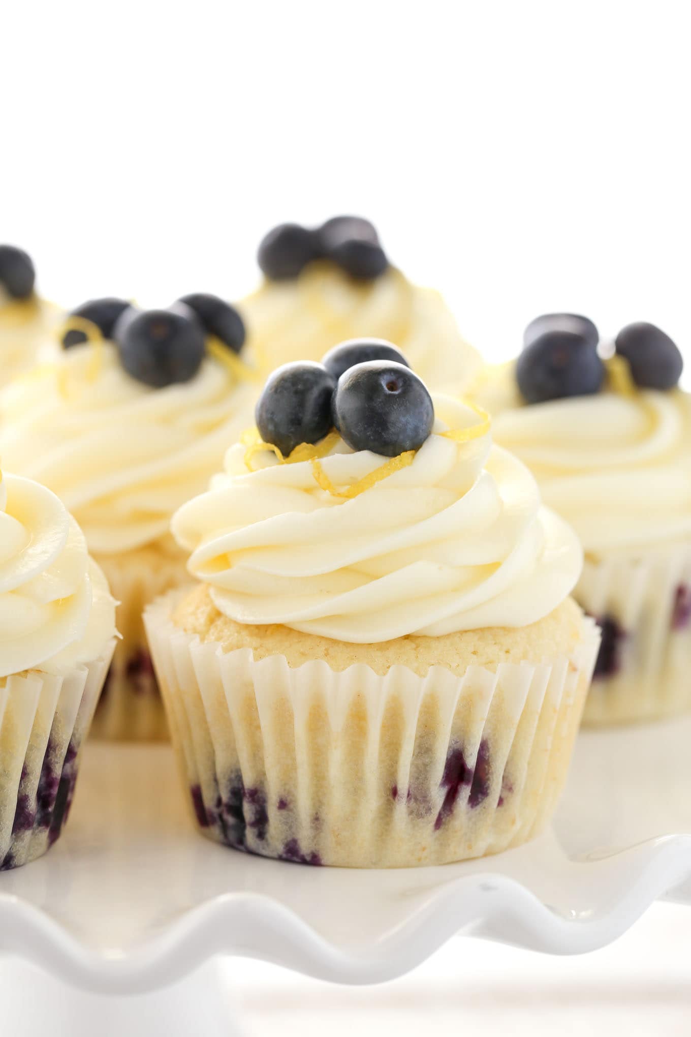 Side view of lemon blueberry cupcakes topped with lemon frosting on a white cake stand. 