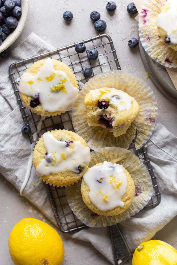 Overhead view of four lemon blueberry muffins on a wire rack. One muffin has a bite missing and is turned on its side. Lemons and fresh blueberries surround the muffins. 