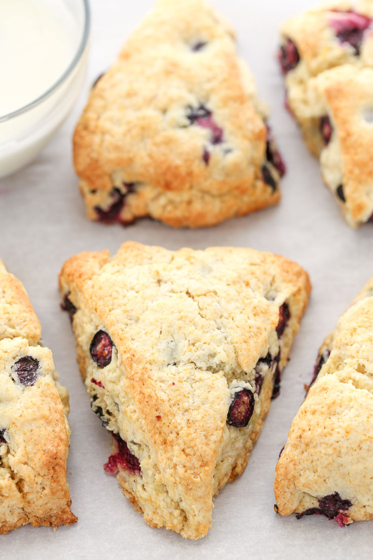 A closeup shot of lemon blueberry scones on parchment paper and a glass bowl filled with lemon glaze.