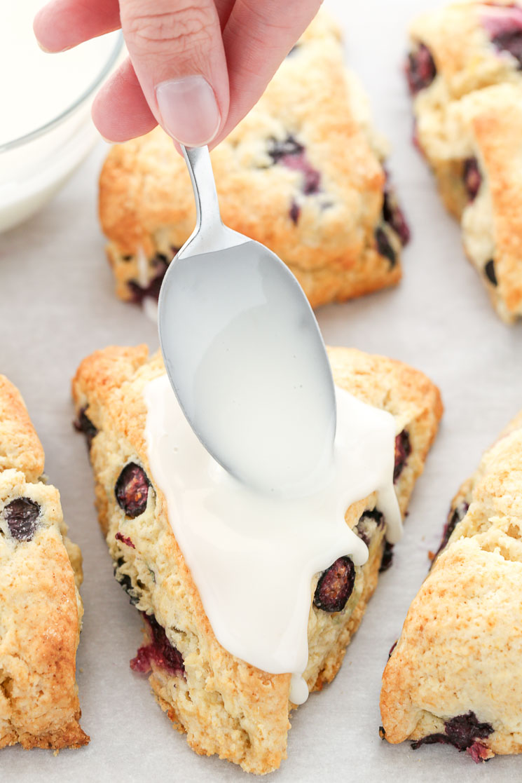 A closeup of the lemon blueberry scones on parchment paper being topped with the lemon glaze.