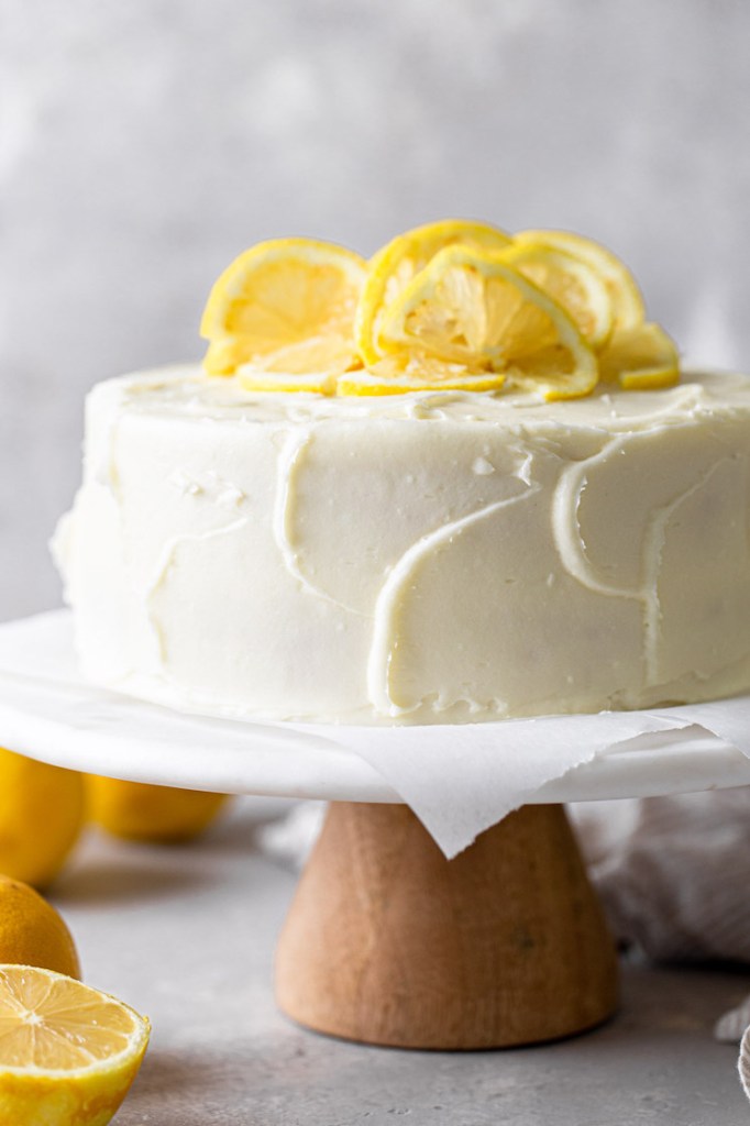 A side view of a lemon layer cake garnished with lemon slices on a white cake stand. 